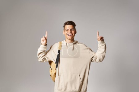 fröhlicher Student in seinen Zwanzigern mit Rucksack, der mit den Fingern nach oben auf grauem Hintergrund zeigt
