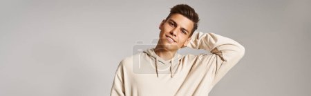 horizontal shot of charming young man with brown hair putting hand behind head on light background
