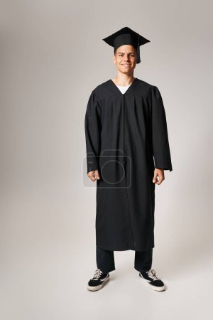 happy attractive student in graduate gown and cap standing against light background