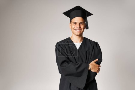 handsome student in graduate gown and cap with grey eyes folded arms against light background