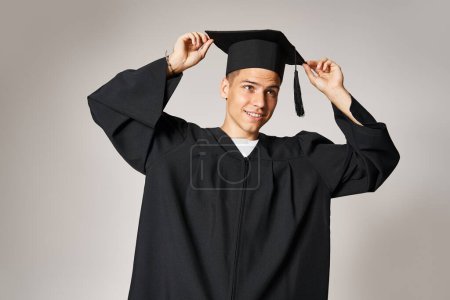 attraktive Studentin im Kleid mit grauen Augen, die vor hellem Hintergrund die Absolventenmütze auf dem Kopf hält