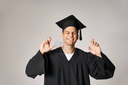 handsome student in graduate gown and cap making wish with closed eyes against grey background