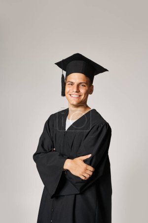 charmant étudiant en robe d'études supérieures et casquette avec les bras croisés souriant et regardant à la caméra
