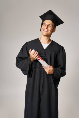 attractive student in graduate gown and cap holding to diploma with hands and looking to forward