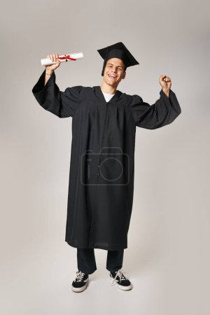 happy handsome student in graduate gown and cap rejoices in receiving diploma on grey background