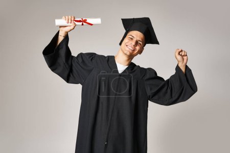 charming young student in graduate gown and cap rejoices in receiving diploma on grey background