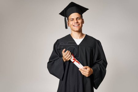 Photo for Portrait of young student in graduate gown and cap holding to diploma with hands on grey background - Royalty Free Image