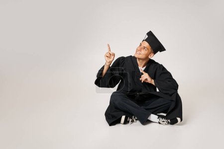 Photo for Cheerful young man in graduate gown and cap sitting and showing with fingers to up - Royalty Free Image