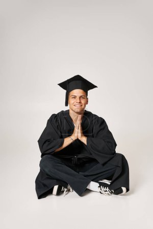 atractivo joven en vestido de graduado y gorra sentado y plegable manos en fondo gris