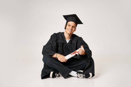 handsome man in graduate gown and cap sitting and holding to diploma with hands on grey background