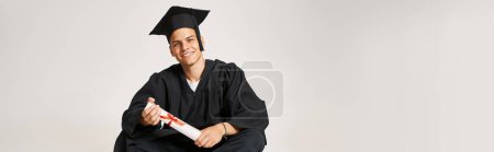 bannière de l'homme heureux en robe d'études supérieures et casquette assis et tenant au diplôme avec les mains