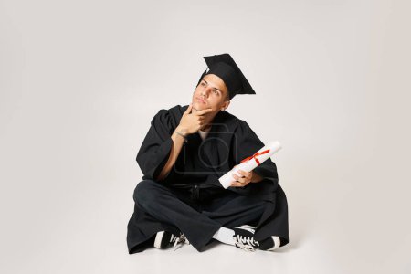 Nachdenklicher Typ im Diplom-Outfit sitzt und hält mit der Hand zum Diplom auf grauem Hintergrund