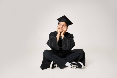 smiling charming guy in graduate outfit sitting and holding with hands to cheeks on grey background