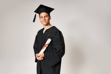 charismatic young student in graduate outfit holding to diploma with hands on grey background