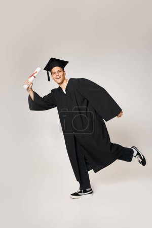 Verspielter junger Mann im Diplom-Outfit posiert mit Diplom in der Hand auf grauem Hintergrund