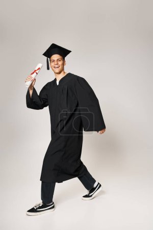 charming student in graduate outfit walking with diploma in hand on grey background