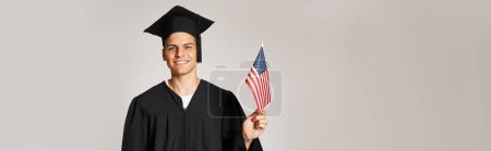 estandarte de estudiante en traje de graduado posando con bandera americana con la mano sobre fondo gris