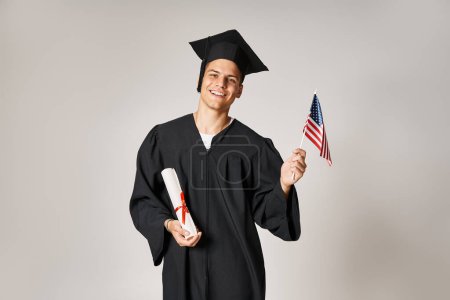 cheerful man in graduate outfit posing with American flag and diploma with hands on grey background