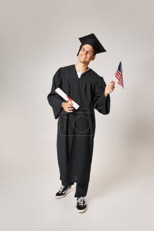 happy student in graduate outfit posing with American flag and diploma with hands on grey background