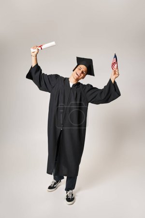 joven estudiante americano en traje de graduado feliz de haber completado sus estudios sobre fondo gris