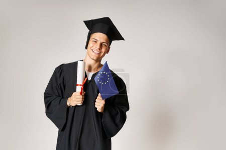 atractivo chico en traje de graduado posando con diploma y bandera europea en manos sobre fondo gris