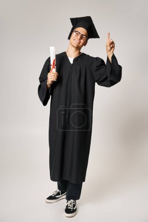 fröhlicher junger Mann im Diplom-Outfit mit Sehbrille, der mit dem Finger nach oben zeigt, Diplom in der Hand