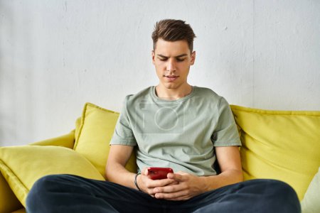 attraktiver Mann mit braunen Haaren zu Hause auf gelbem Sofa sitzend und SMS in Smartphone