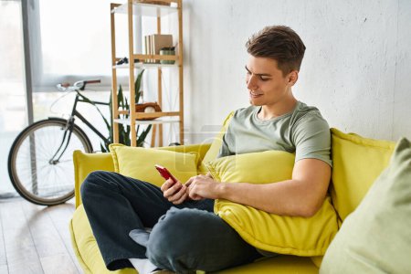 lächelnder Typ in seinen Zwanzigern zu Hause auf gelbem Sofa sitzend und in den sozialen Medien scrollend