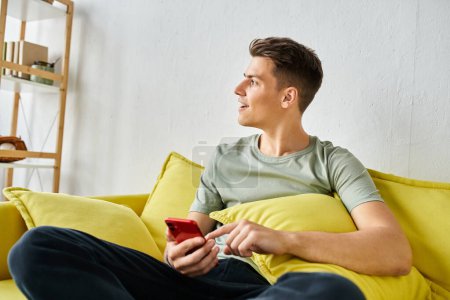 Charmanter Typ in seinen Zwanzigern zu Hause sitzt auf der gelben Couch in den sozialen Medien und schaut zum Fenster