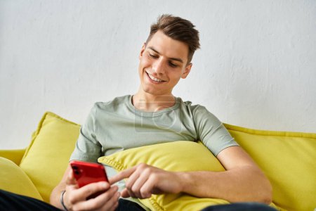 cheerful young man with brown hair at home sitting on yellow couch and scrolling in social media