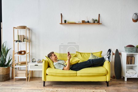 Téléchargez les photos : Attrayant jeune homme couché sur le canapé jaune dans son salon et défilant vers les médias sociaux - en image libre de droit