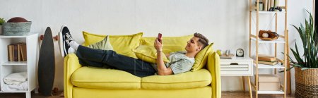 horizontal shot of young man lying on yellow couch in living room and scrolling to social media