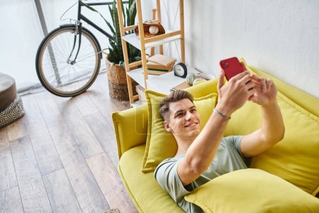 attraktiver junger Mann mit braunen Haaren liegt auf gelbem Sofa im Wohnzimmer und macht Selfie