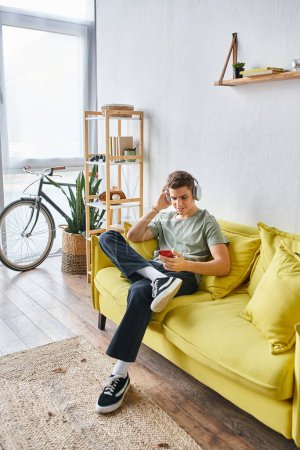 schöner junger Mann mit Kopfhörern, der zu Hause auf einer gelben Couch sitzt und in den sozialen Medien blättert