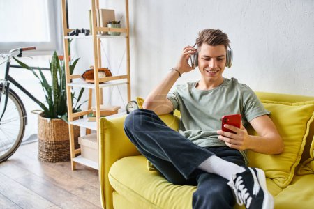Téléchargez les photos : Jeune homme souriant dans les écouteurs assis sur le canapé jaune à la maison et faisant défiler vers les médias sociaux - en image libre de droit