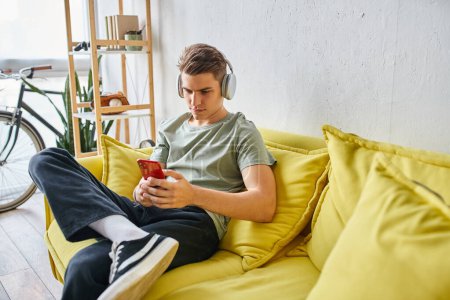 Junge Studentin sitzt mit Kopfhörer auf gelbem Sofa zu Hause und textet auf Smartphone