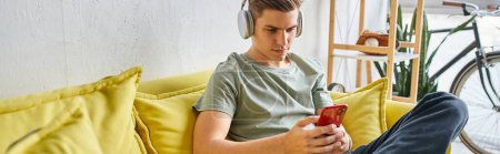 horizontal shot of student in headphones sitting on yellow couch at home and texting on smartphone