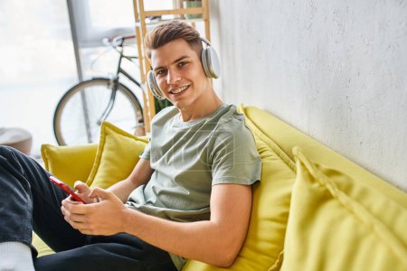 Téléchargez les photos : Homme souriant dans les écouteurs dans le canapé jaune à la maison textos sur smartphone et regardant à la caméra - en image libre de droit
