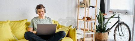 Foto de Estandarte de joven atractivo en auriculares en sofá amarillo en casa estudiando en el ordenador portátil - Imagen libre de derechos