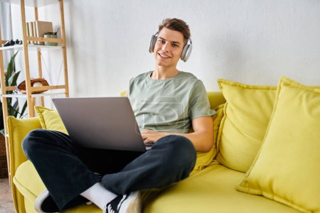 Foto de Joven guapo con auriculares y portátil en sofá amarillo en casa mirando a la cámara - Imagen libre de derechos
