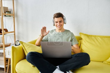 cheerful student with headphones and laptop in yellow couch saying hello to online meeting