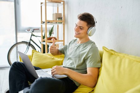 smiling young man with headphones and laptop in yellow couch liking to online meeting