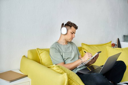 Téléchargez les photos : Étudiant concentré avec écouteurs et ordinateur portable dans le canapé jaune étudier et écrire dans la note - en image libre de droit