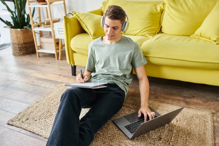 student with headphones on floor near yellow couch studying in laptop and writing in note