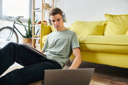 focused young man with headphones on floor near yellow couch studying in laptop and writing in note