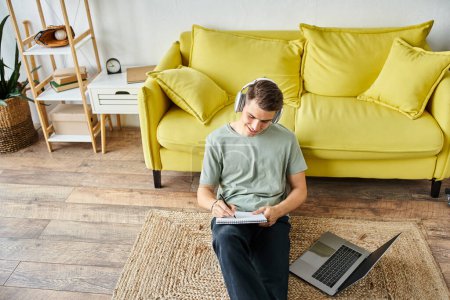 smiling guy with headphones and laptop on floor near yellow couch studying and writing in note