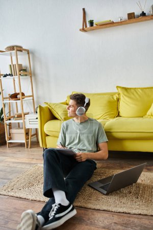 guy with headphones and laptop on floor near yellow couch writing in note and looking to window