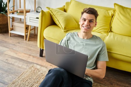 Charmanter junger Mann auf dem Boden in der Nähe der gelben Couch zu Hause sitzt mit Laptop und lächelt in die Kamera