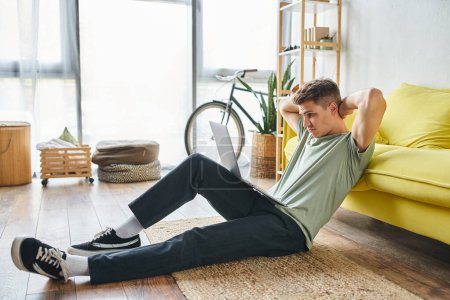 Foto de Hombre confundido en el suelo cerca de sofá amarillo en casa estudiando en el ordenador portátil y poniendo las manos detrás de la cabeza - Imagen libre de derechos