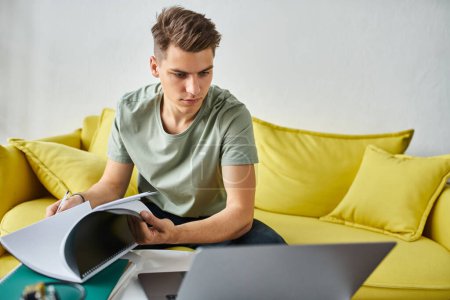 young man in yellow couch concentration doing coursework with notes and laptop on coffee table